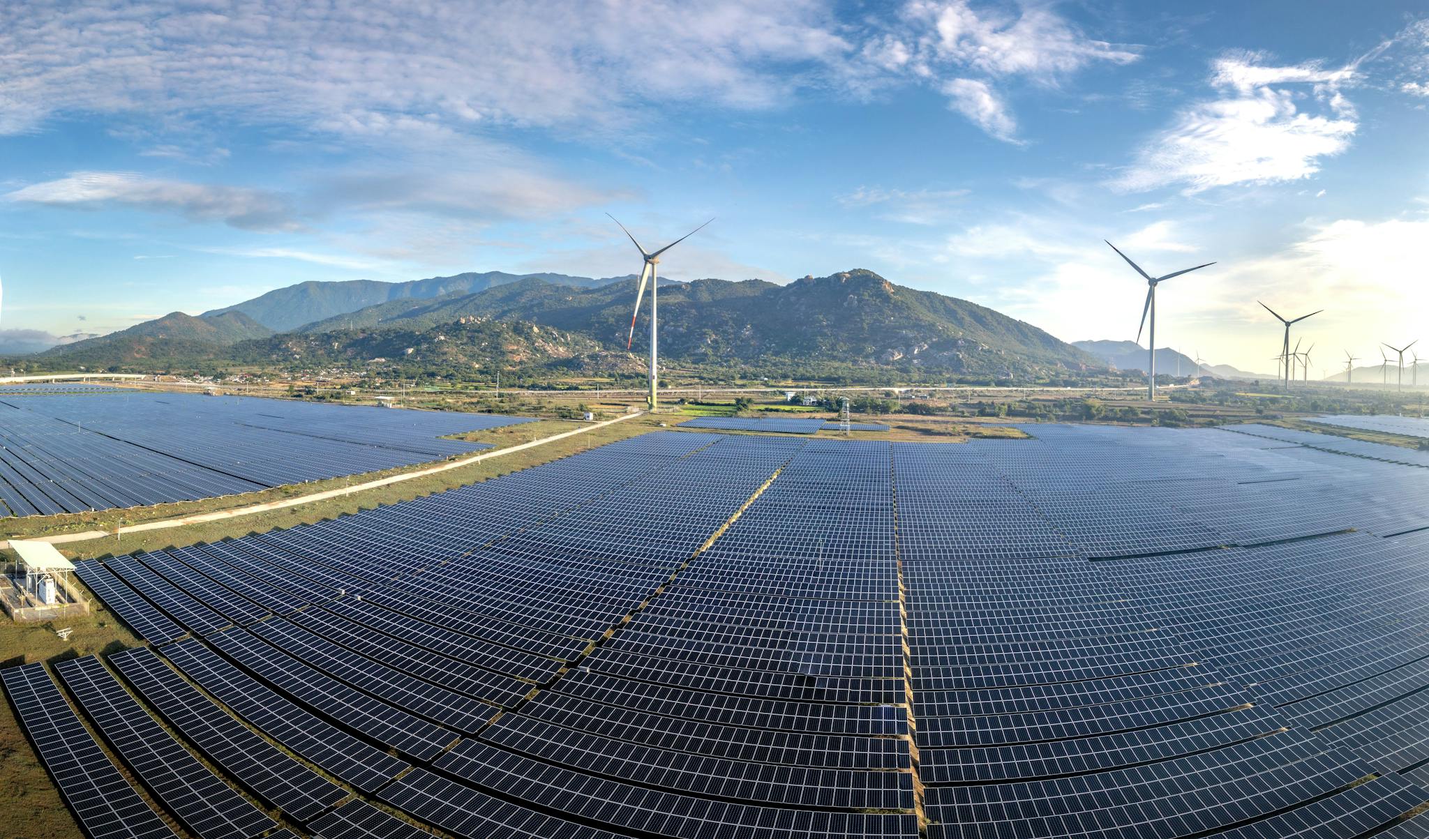 Aerial view of a vast solar farm with wind turbines against a mountainous backdrop on a sunny day.